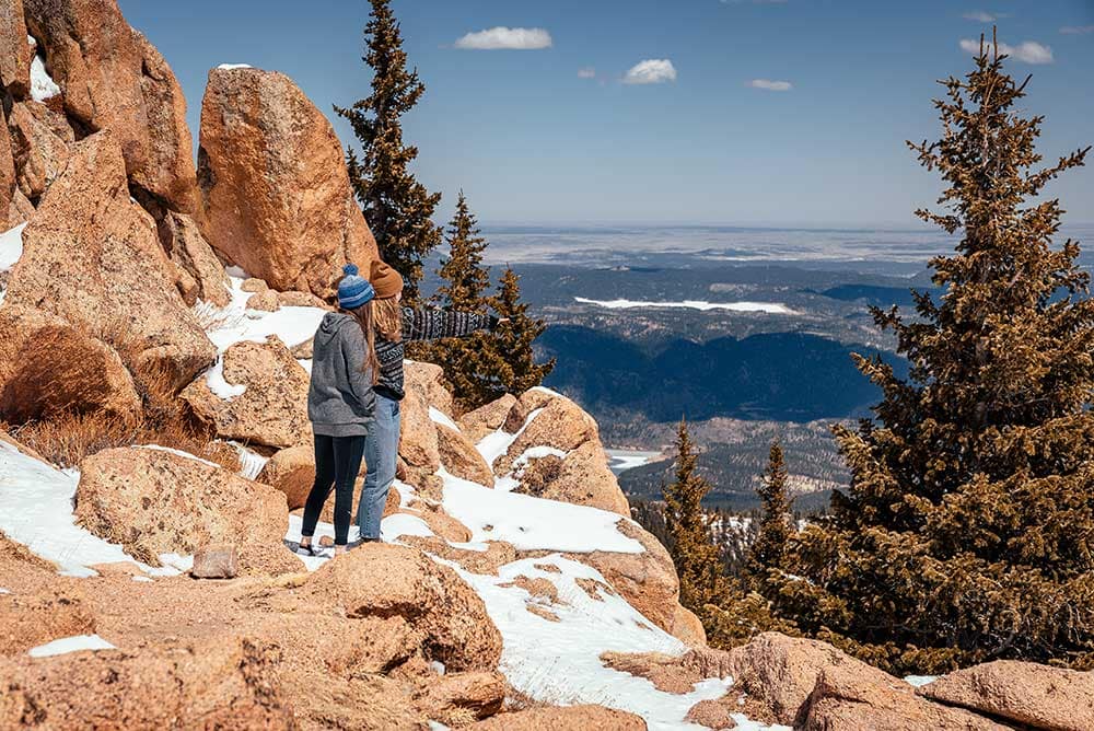 Two women hike along red rocks overlooking the mountains below. 