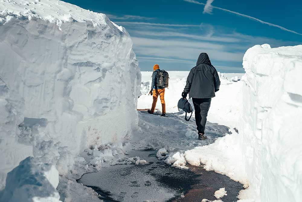 Two hikers weave amongst towering walls of snow dressed in warm weather gear. 