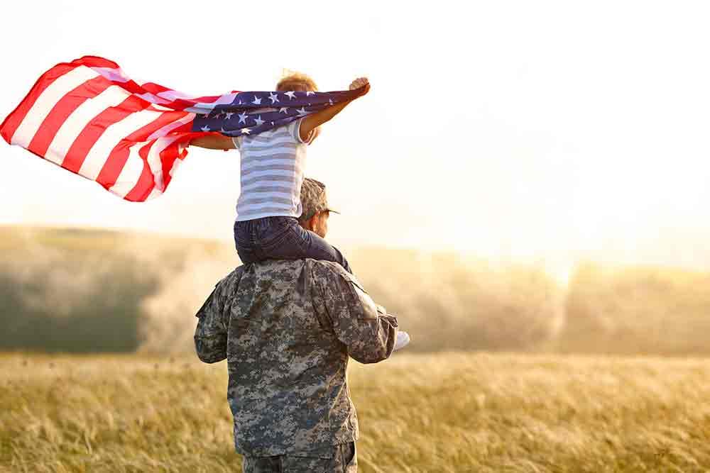 Military service member holding a child on their back with the American flag.
