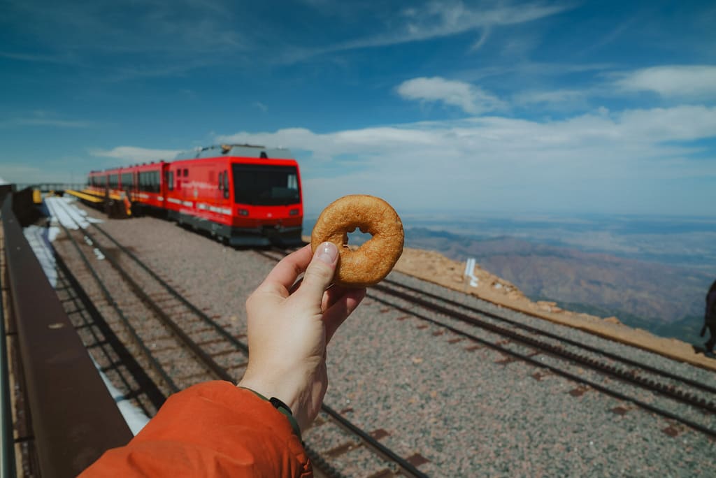 Person holding donut on top of Pikes Peak with the Pikes Peak Cog Railway in the background