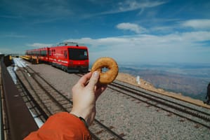 Person holding donut on top of Pikes Peak with the Pikes Peak Cog Railway in the background